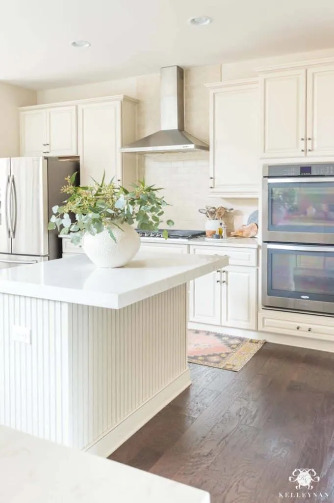 A cream kitchen with a double wall oven and large hood vent with a white vase of green eucalyptus on the island.