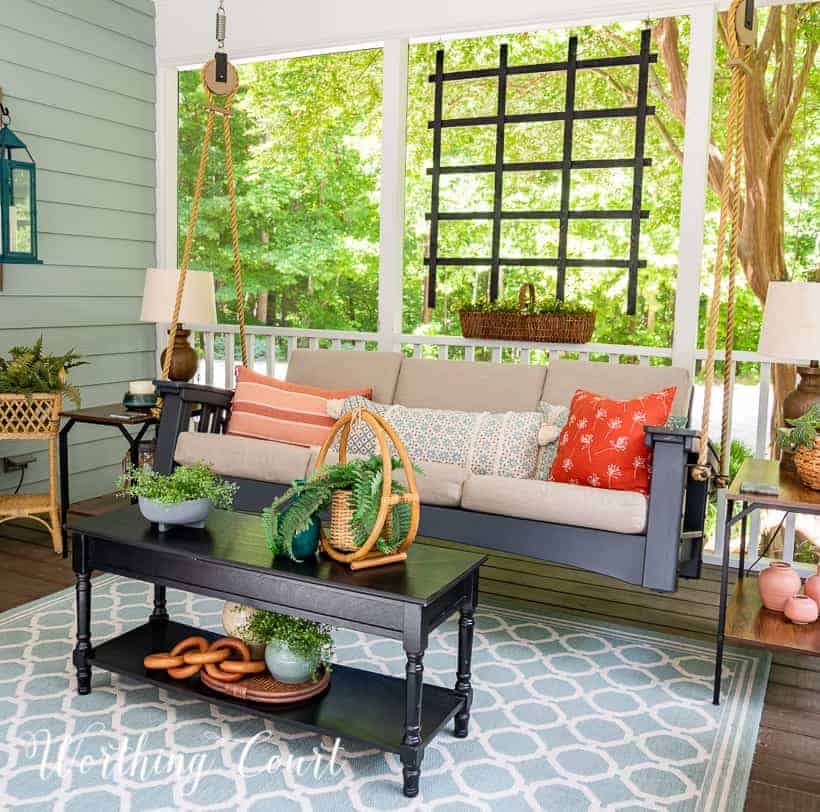 A screened in porch with a hanging swing, a wood coffee table and a blue and white rug.