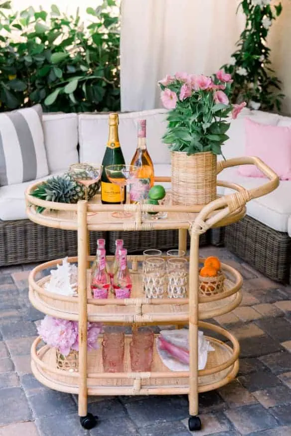 A light wood bar cart with champagne bottles, glasses and a plant with pink flowers.