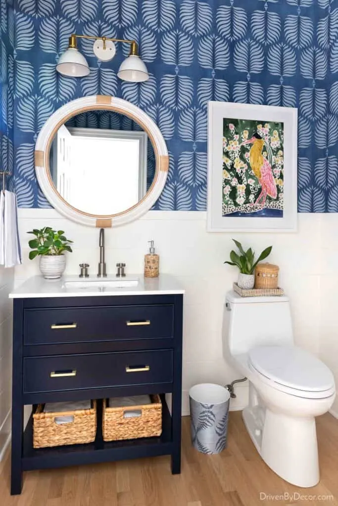 White shiplap with blue and white wallpaper above in a bathroom with a navy cabinet, white counter and round white and tan mirror.