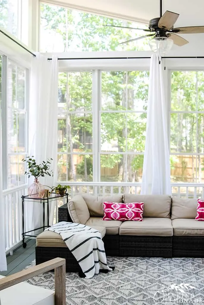 A sectional sofa with beige cushions on a screened in porch with white sheer curtains.