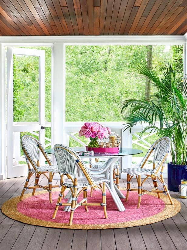 A glass round table and blue and white wicker chairs with a pink and yellow rug under.  A wood ceiling and large palm tree in the corner.