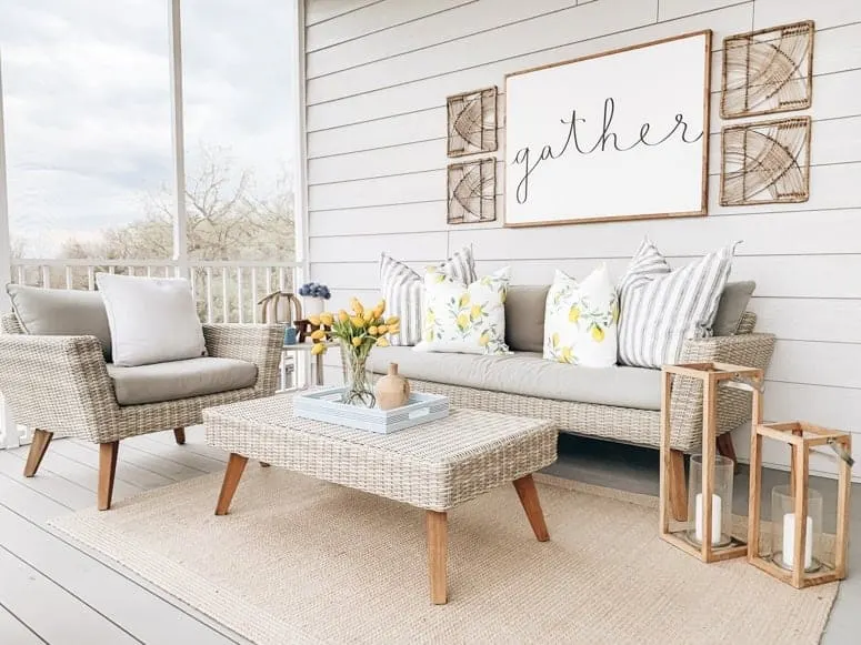A screened in porch with neutral furniture, a neutral colored rug, two lanterns and a sign on the wall that says gather.