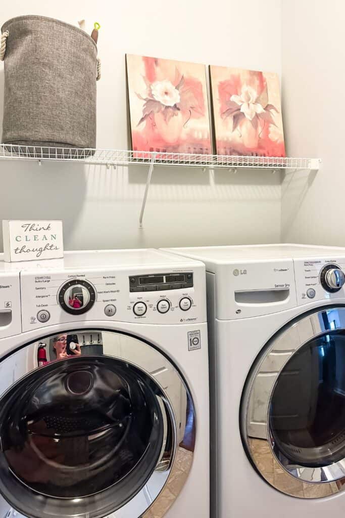 White washer and dryer with a wire shelf above and two floral prints, a gray laundry basket.