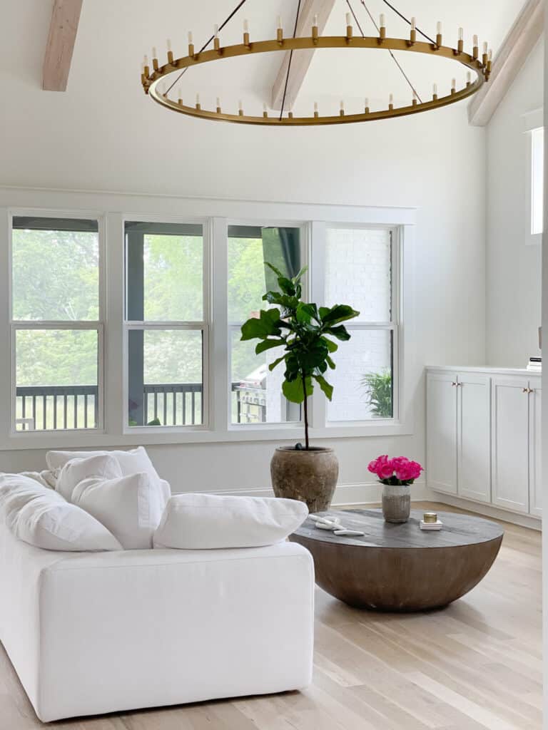 A family room with lots of windows, a large wagon wheel chandelier, white cabinets with gold knobs, a whtie sofa, light colored wood floors and a round wood coffee table.