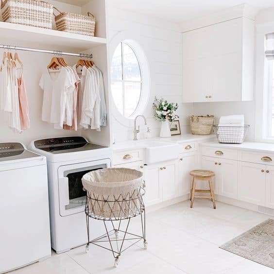 A big white laundry room with a hanging bar above the machines, a large rolling laundry basket and a wood stool in the corner.