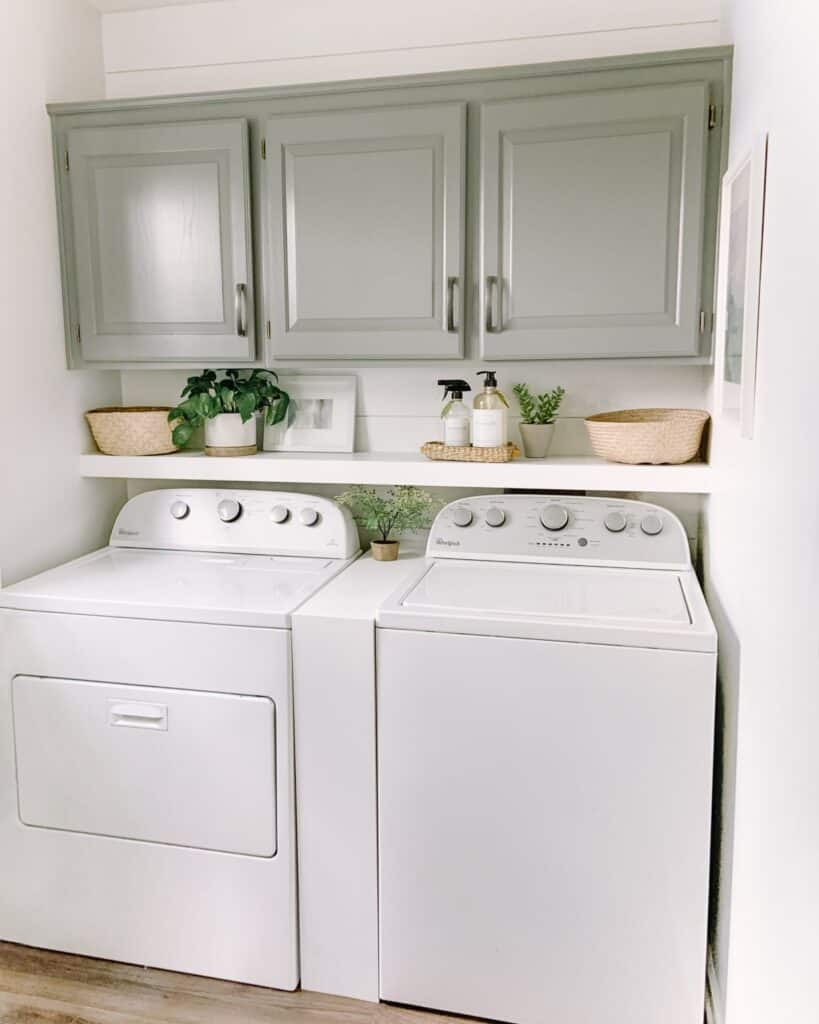 A laundry room with soft gray painted cabinets, a shelf above the machines and a shelf in between them.