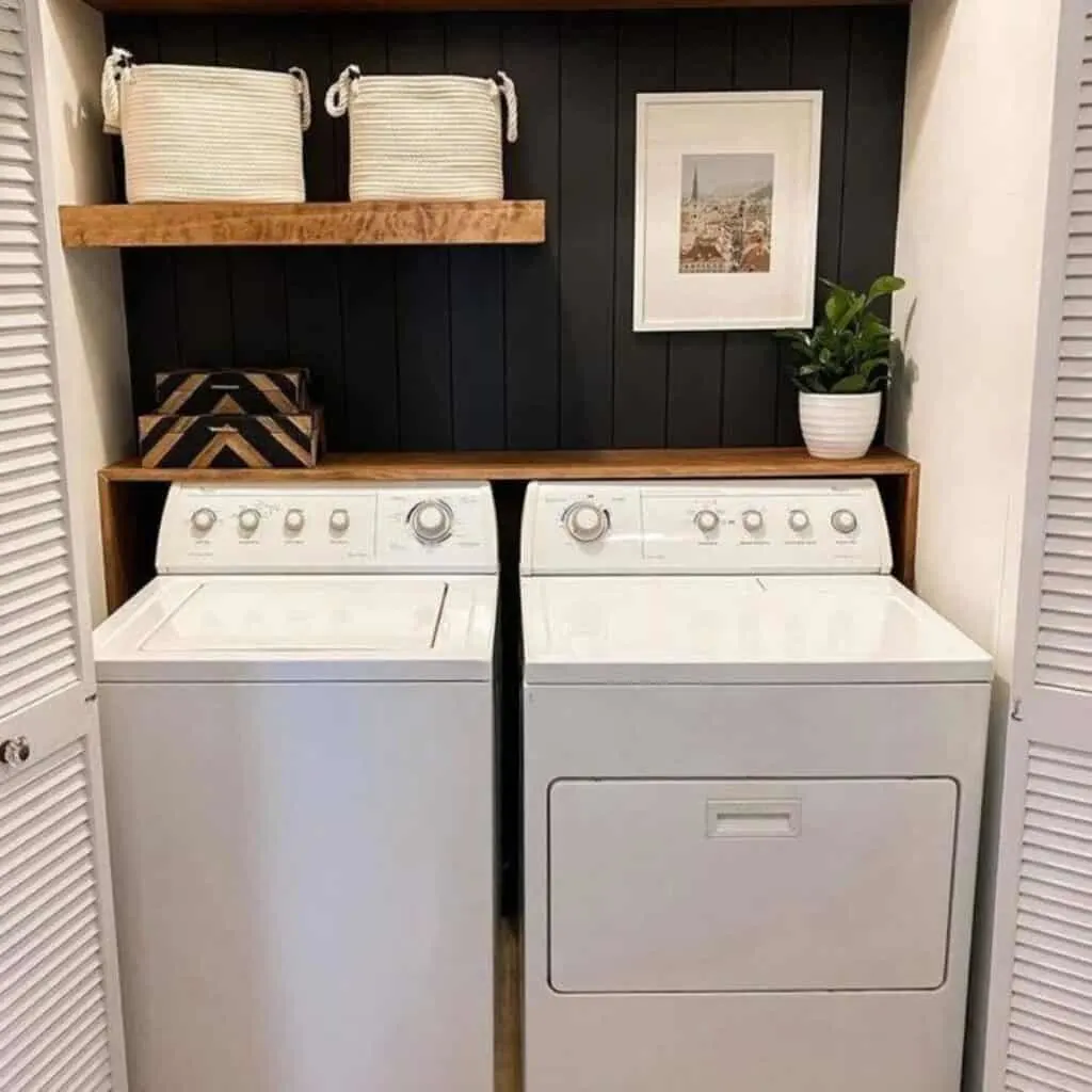 A laundry room with a vertical black shiplap on the back wall and wood shelves white baskets and a landscape picture.