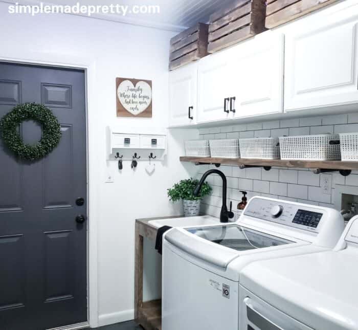 A white laundry room with subway tile backsplash and accents of wood on the sink base, shelf and crates above the cabinets.