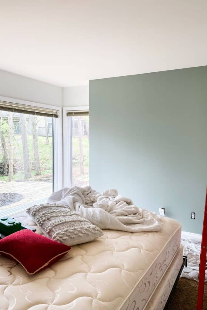 A wall painted sage with an unmade bed and large windows.