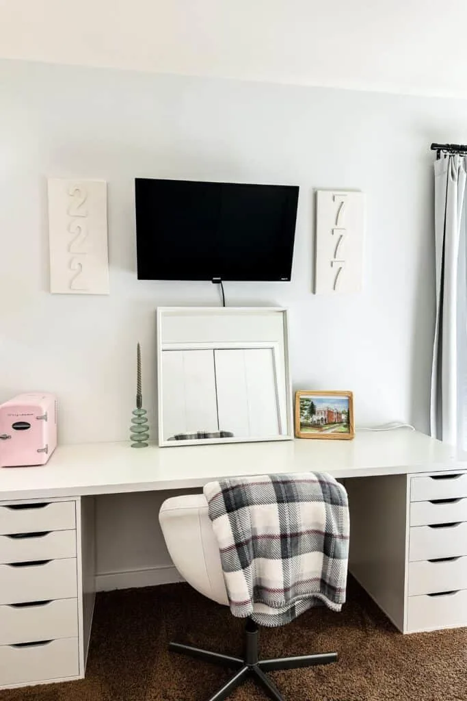 A desk with a leaning mirror, wall mounted tv and a pink tiny fridgte on the desk.