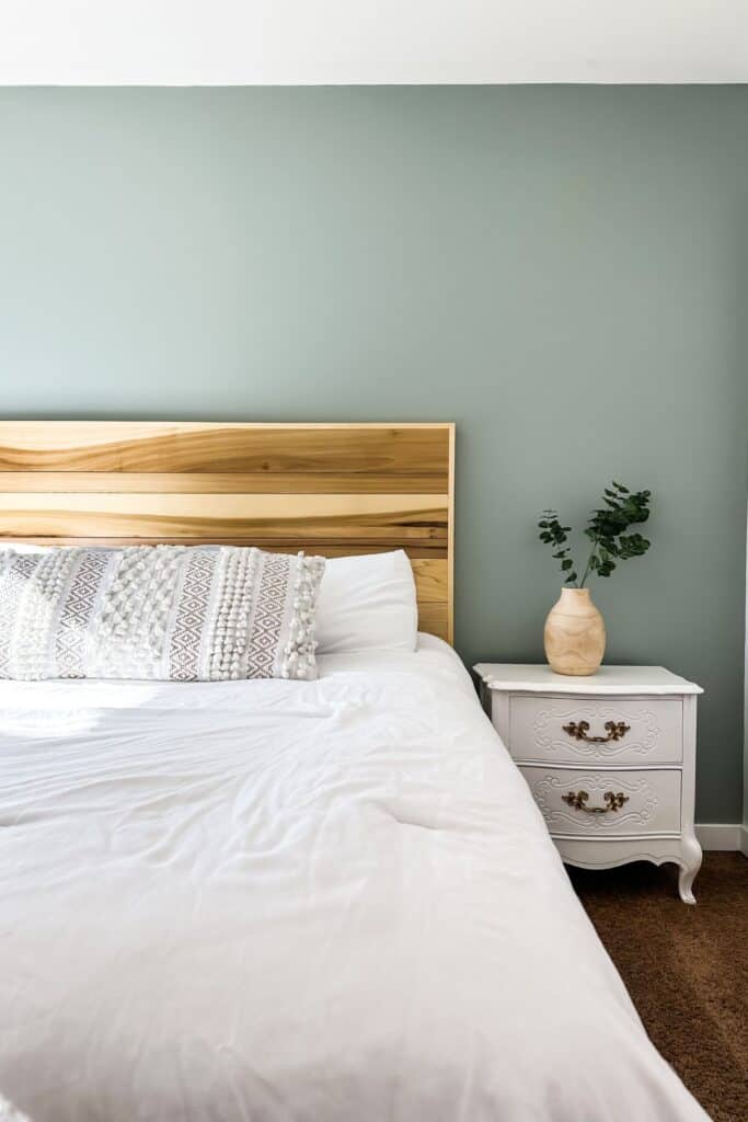A wood headboard against a sage green wall, white bedding and a white nightstand.