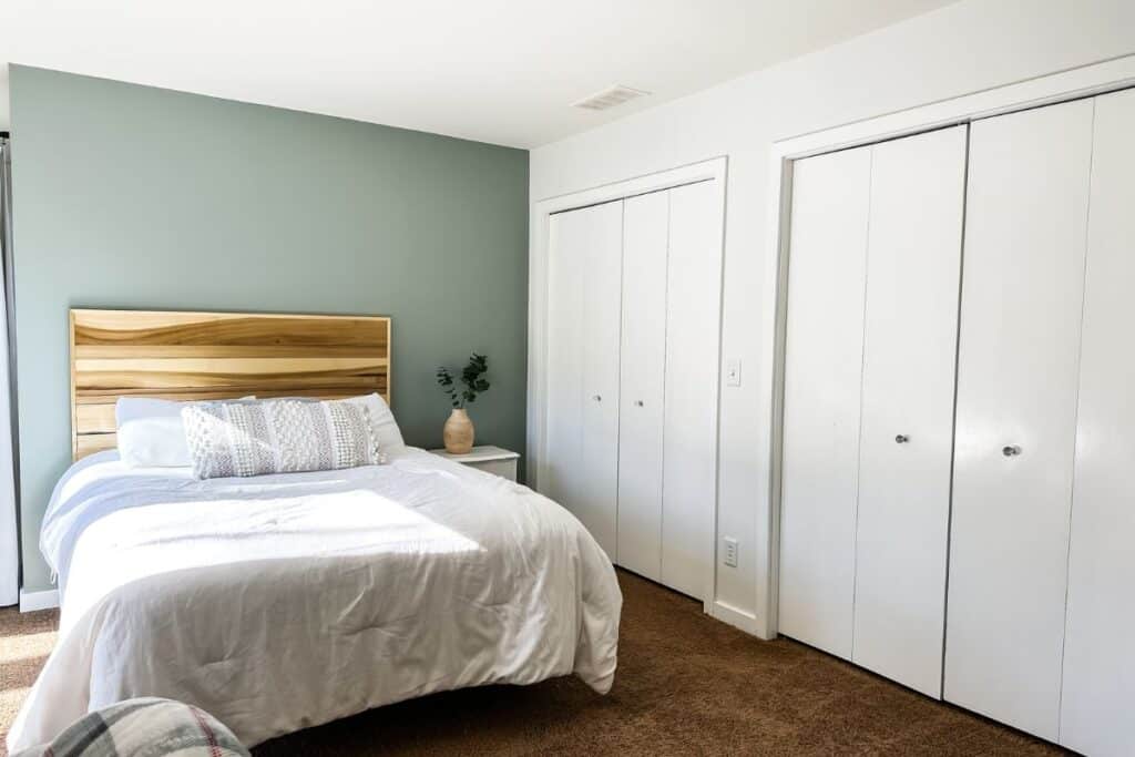 A bedroom with three white walls and the headboard wall in sage green, a wood headboard and white bedding.