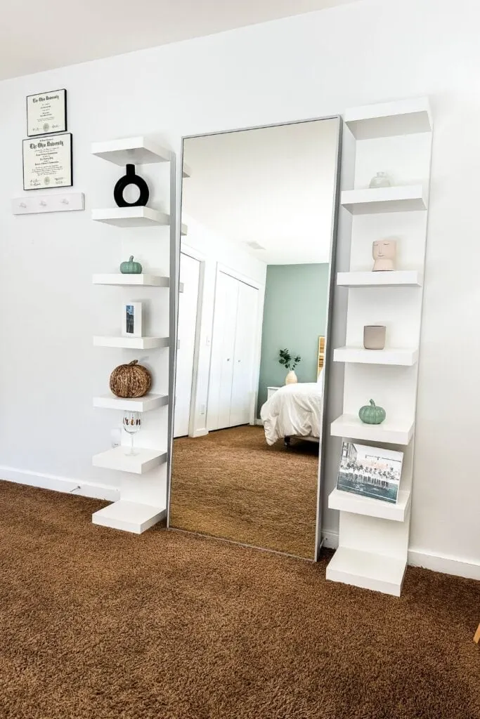 A large mirror leaning on a wall with a set of shelves on either side and brown carpet.