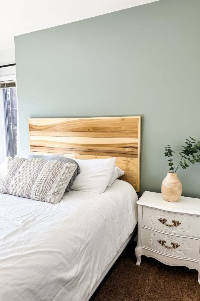 A wood headboard, sage green wall, white bedding and a white nightstand.
