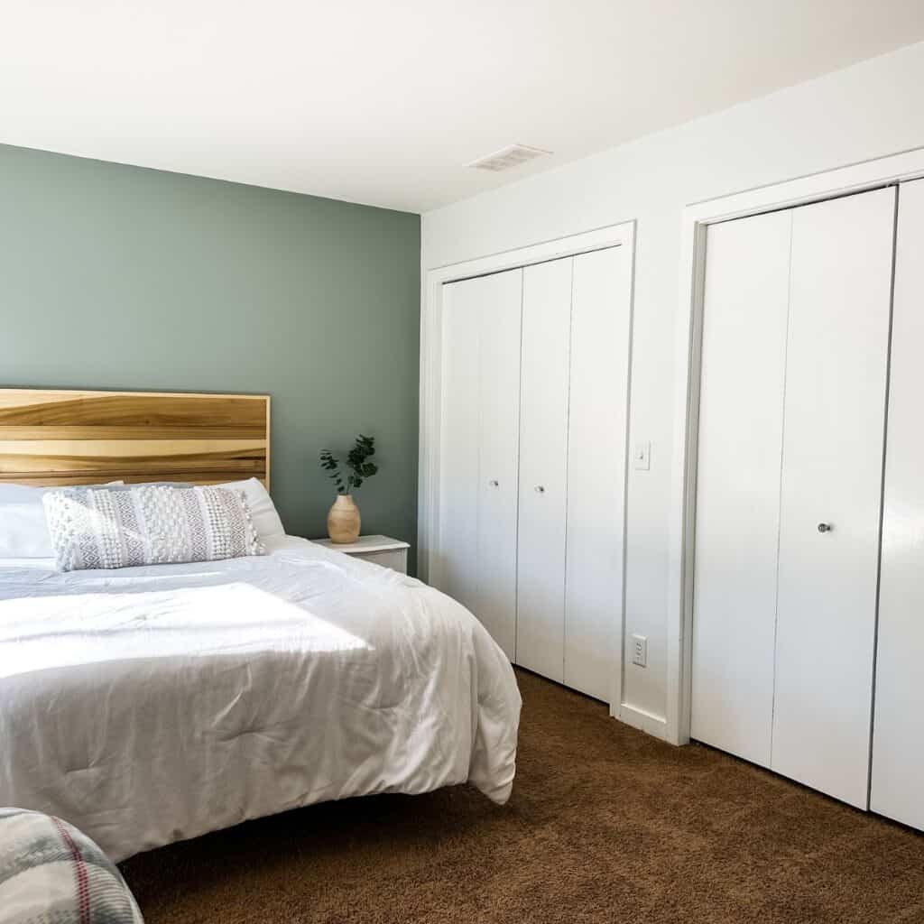 A bedroom with a sage green headboard wall and white walls everywhere else, a wood headboard, white bedding and brown carpet.