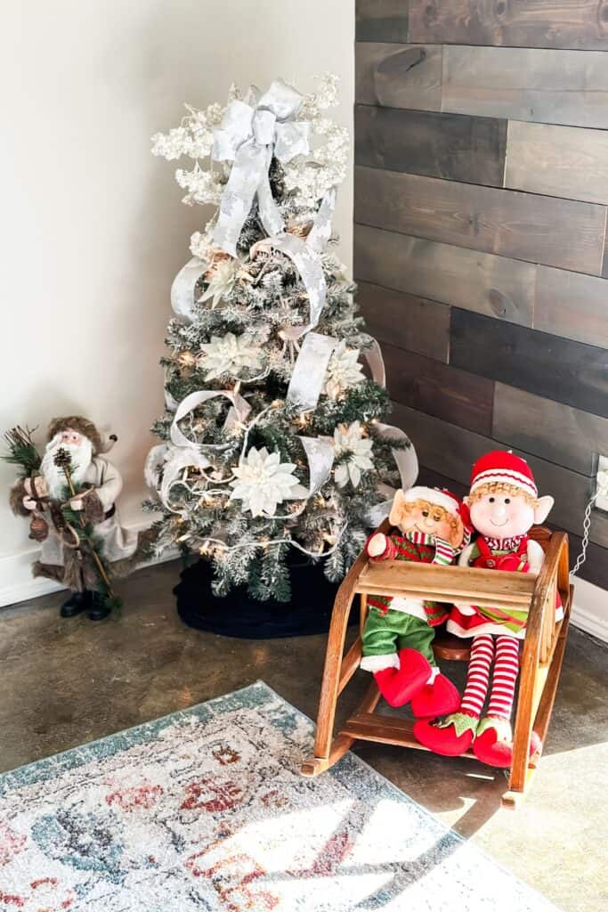 A small flocked Christmas tree with a santa on the left and a childs rocker with two elf dolls in it.