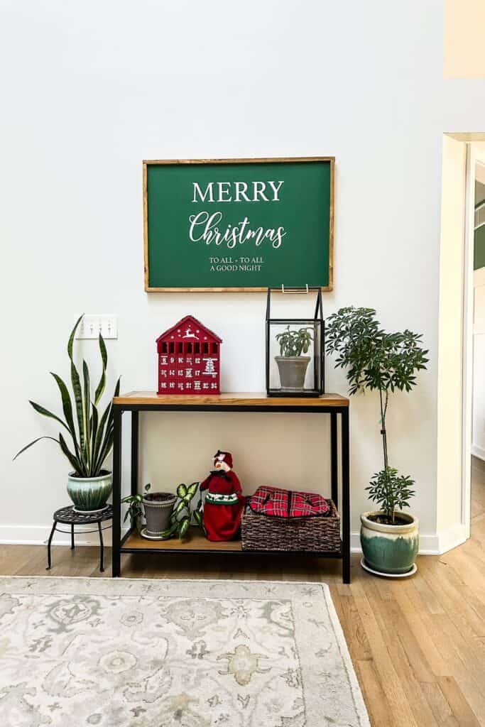 An entryway table with a green merry christmas sign above, a red advent calender in the shape of a house and a red and green female snowman on the bottom shelf next to plants.