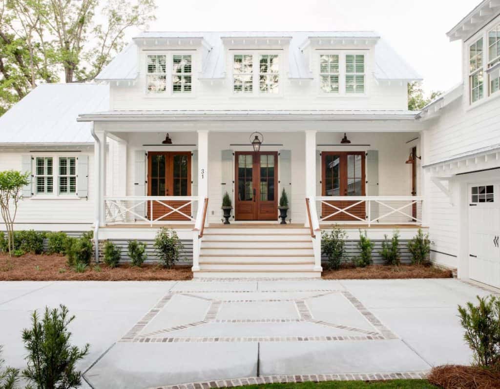 A white house with white trim and a white metal roof with warm wood doors and black light fixtures above. The driveway is white concrete with a design in it made from bricks in a square inside a square.