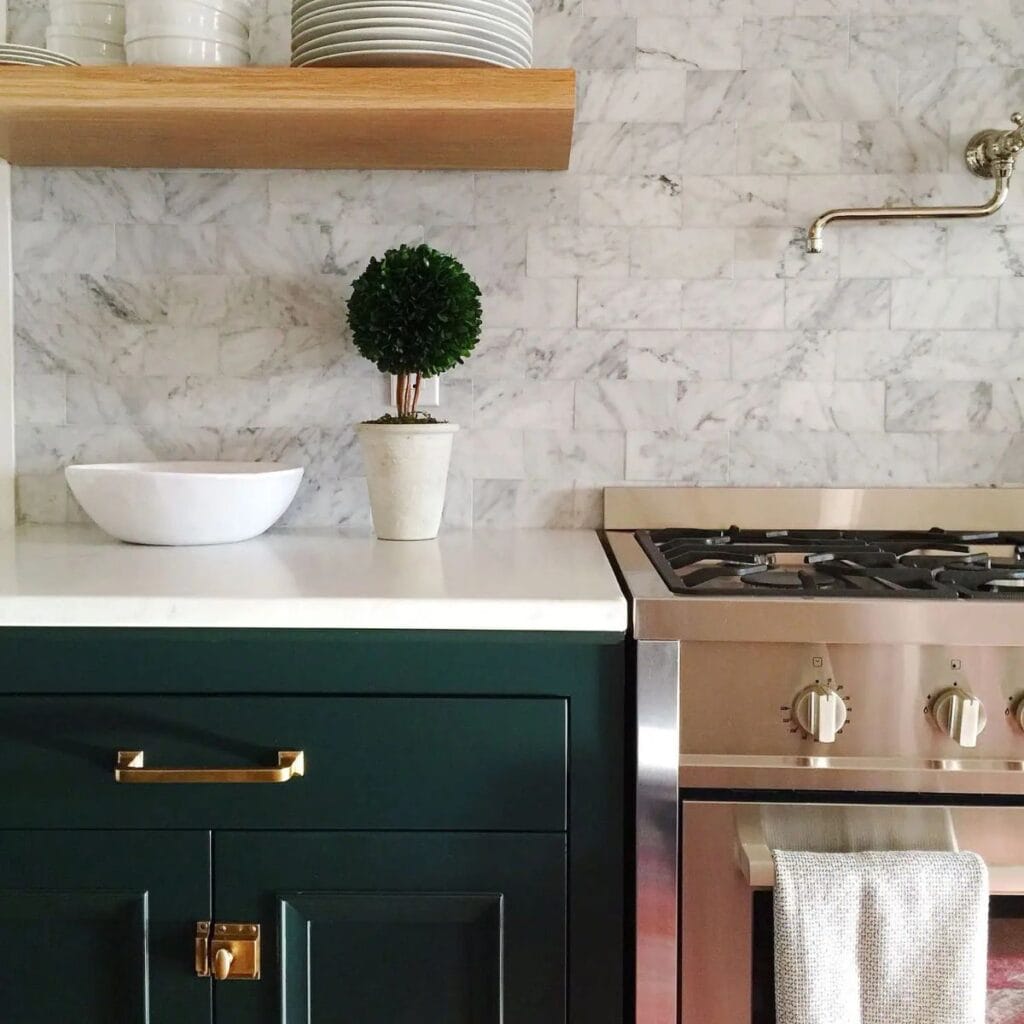 A kitchen with dark green cabinets, gold hardware, a stainless oven, white countertops and marble brick pattern backsplash.