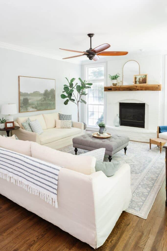 A family room with a painted white brick fireplace and light wood mantel.  It has a wood ceiling fan, light beige couches, light gray tufted ottoman and large artwork on the wall above the sofa.