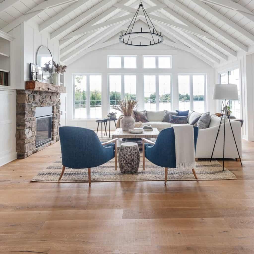 A family room with a stone fireplace, white shiplap wall with exposed beams and a wall of windows overlooking the lake.
