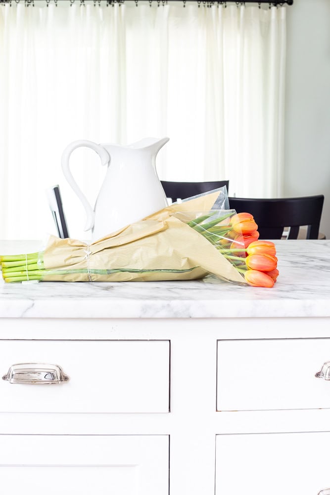 Tulips laying in the store wrapping on a countertop that is marble and a white pitcher next to the tulips.