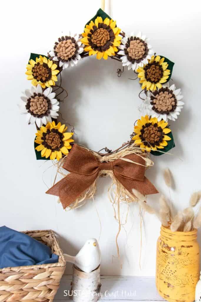 A crafted wreath with yellow and white sunflowers and a bow at the bottom hanging on a white wall.