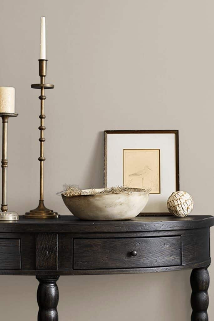 A dark wood desk against a wall painted with Amazing Gray and a wooden bowl candlesticks, a framed piece of artwork of a bird.