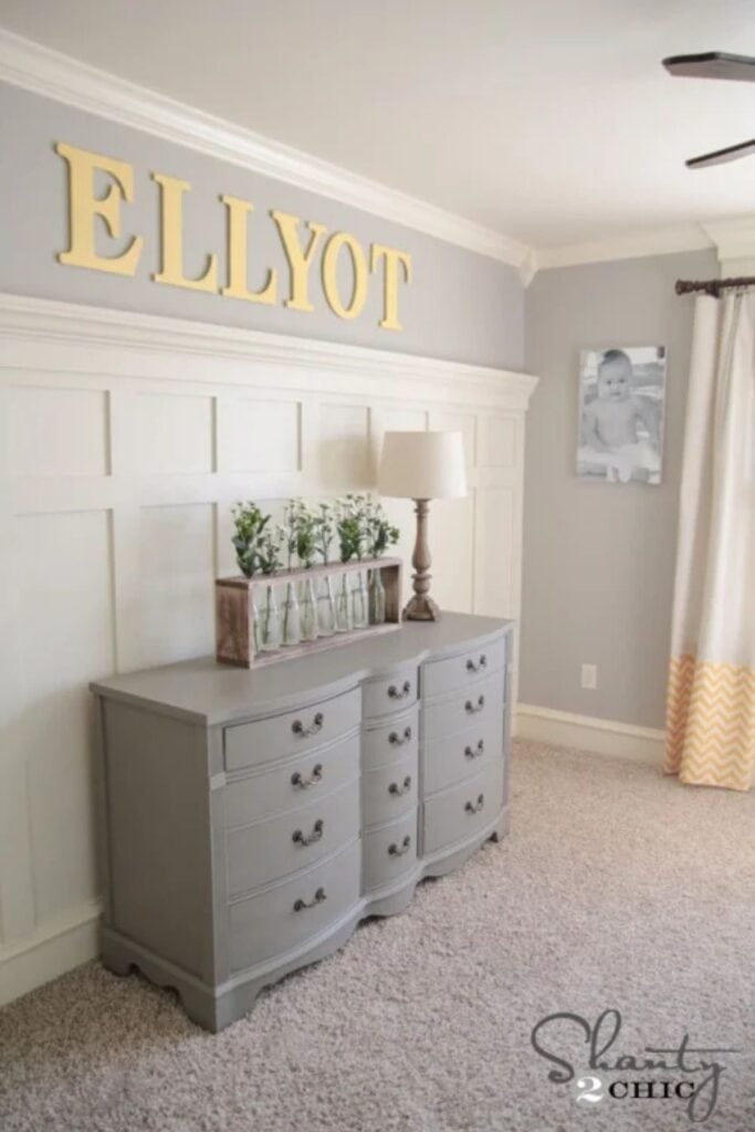 A nursery painted with Amazing Gray and board and batten with a dreser, lamp, and vases of eucalyptus.