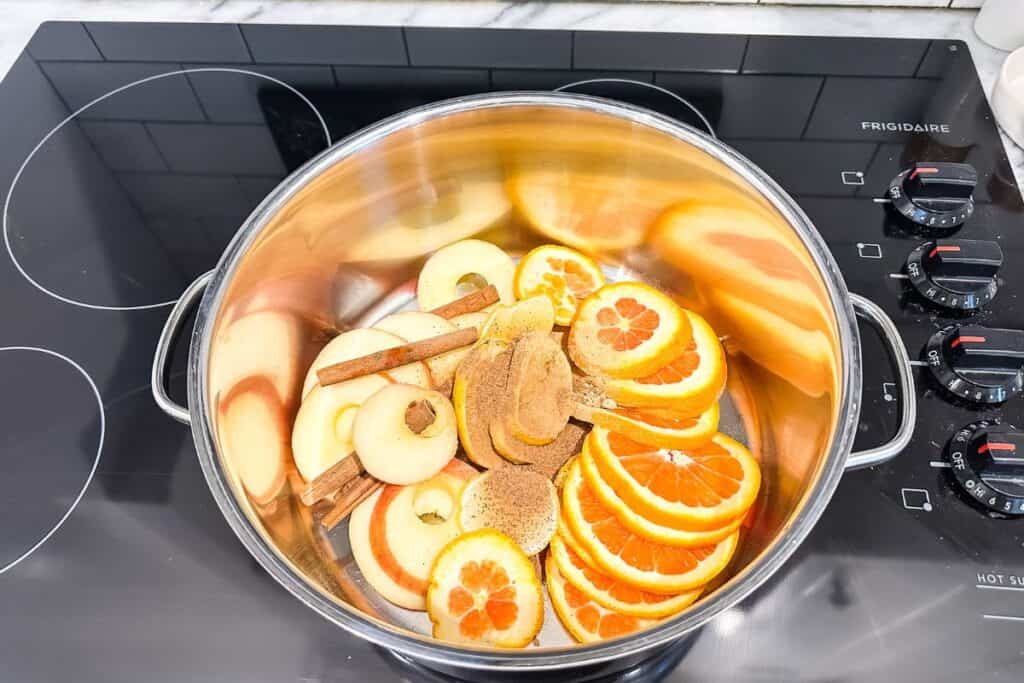 Sliced apple, lemon, and orange with cinnamon sticks and spices in a large pot on a stove.
