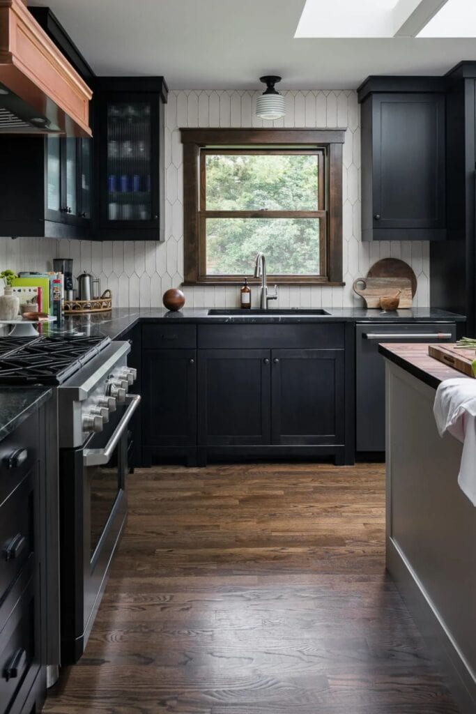 A kitchen with black cabinets, white backsplash, silver hardware, and wood floors.