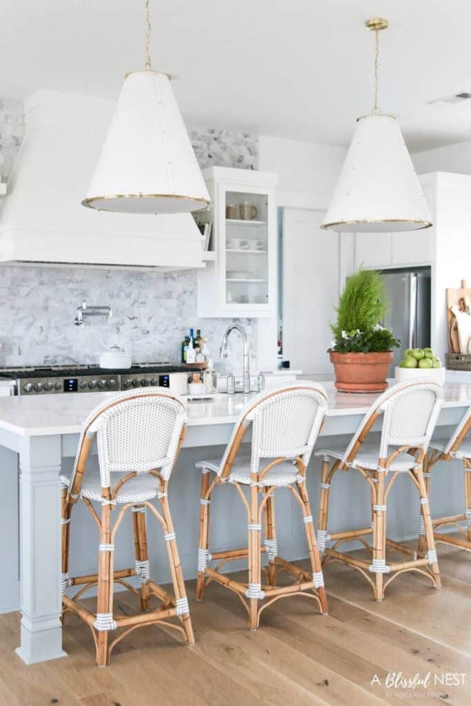A kitchen with light blue cabinets, a marble counter and backsplash with silver hardware, a white oven hood and white light fixtures.