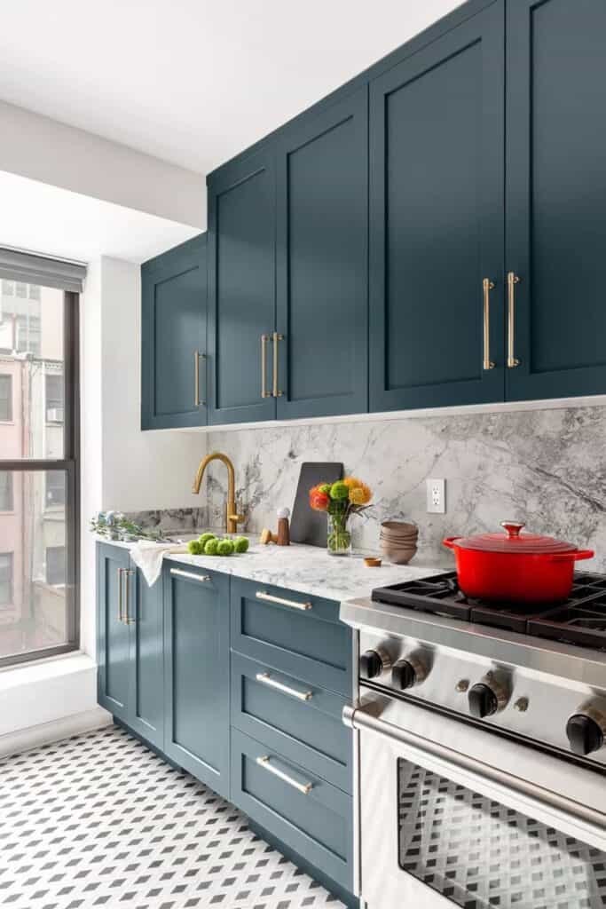 A kitchen with blue cabinets and gold hardware, granite counters and backsplash and a red dutch oven on the stove.