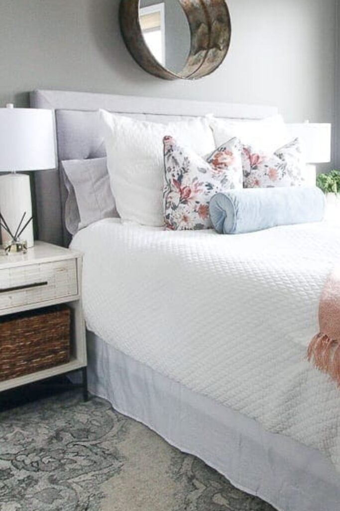 A bedroom with Chelsea Gray on the walls, an upholstered headboard and white bedding with pink throw blanket and floral pillows.