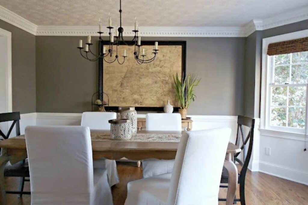 A dining room with the bottom third of the walls painted white under a chair rail, the top painted in Chelsea Gray with wood floors and a black iron chandelier.