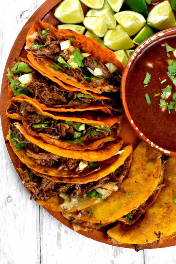Birria Tacos on a round platter with a bowl in the middle with a sauce and slices of limes.