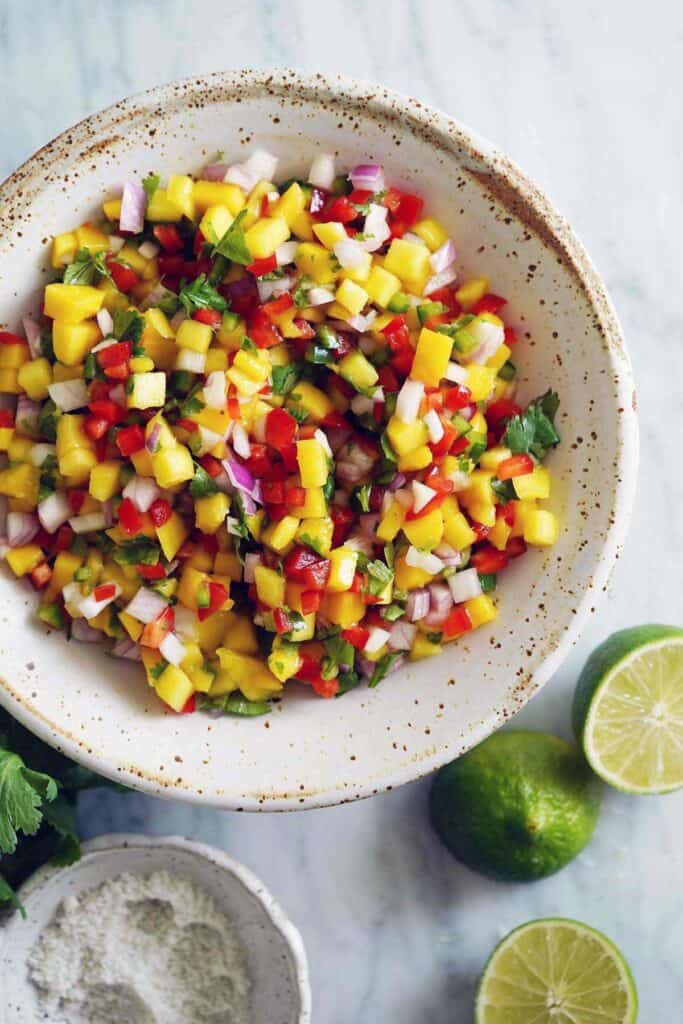 Mango salsa in a white bowl with speckles on it and lime wedges.