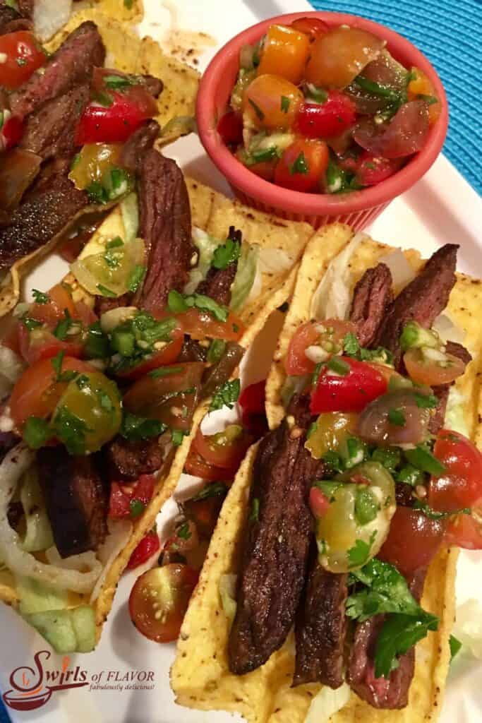 Skirt steak tacos on a white plate with a bowl of pico de gayo in a red bowl.