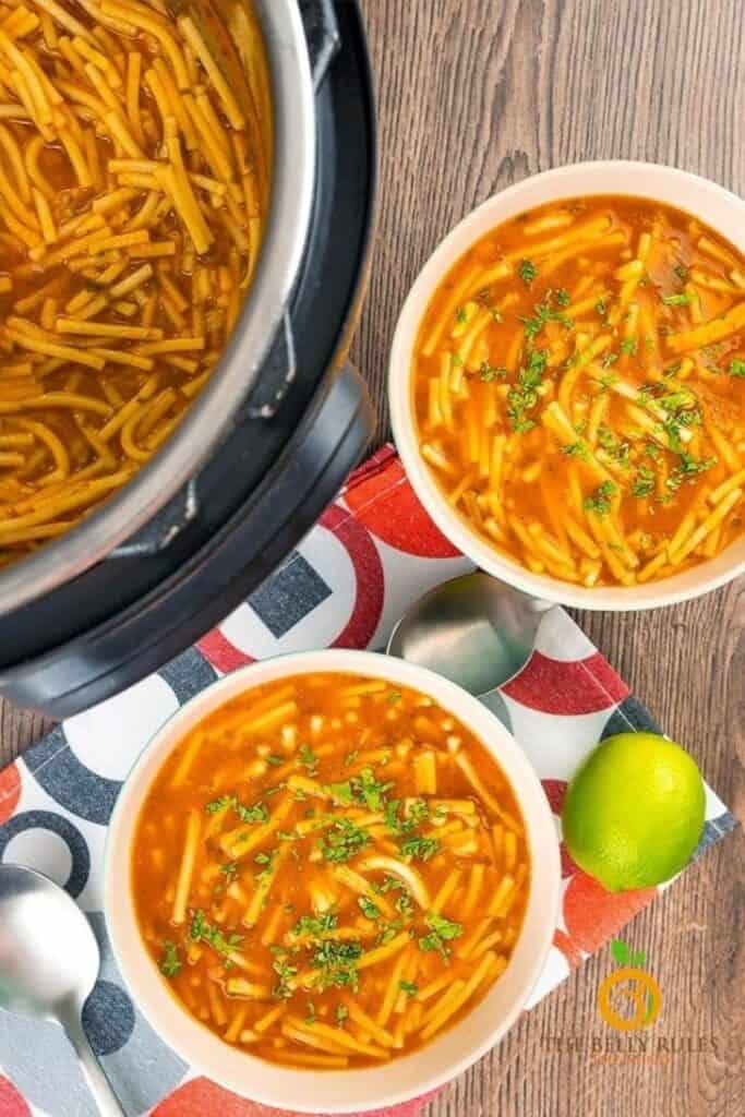 Mexican noodle soup in a crock pot and two white bowls on a wood surface and a spoon and lime.