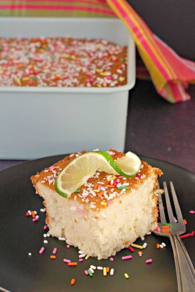 Lime coconut tres leches cake with sprinkles and a lime wedge on a black plate with a fork.