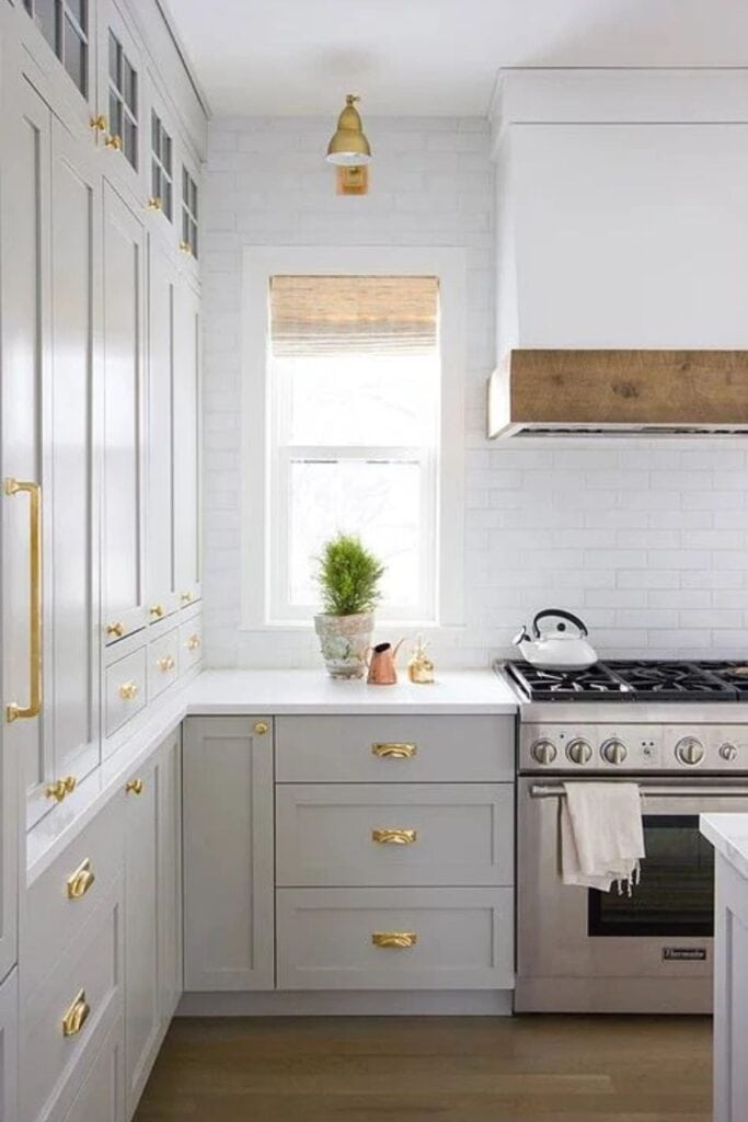 A kitchen with light gray cabinets with gold hardware, a white subway tile backsplash, stainless steel appliances and a small window.