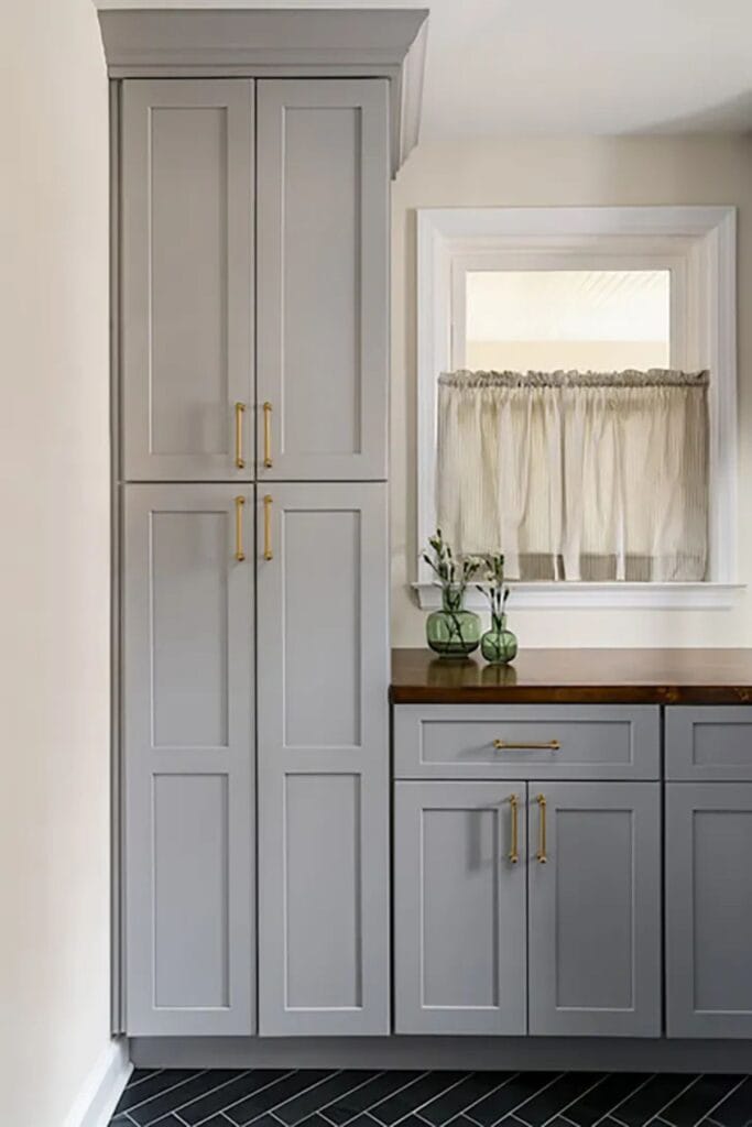 Gray cabinets with gold hardware and wood counters with a window that has a cafe length curtain.