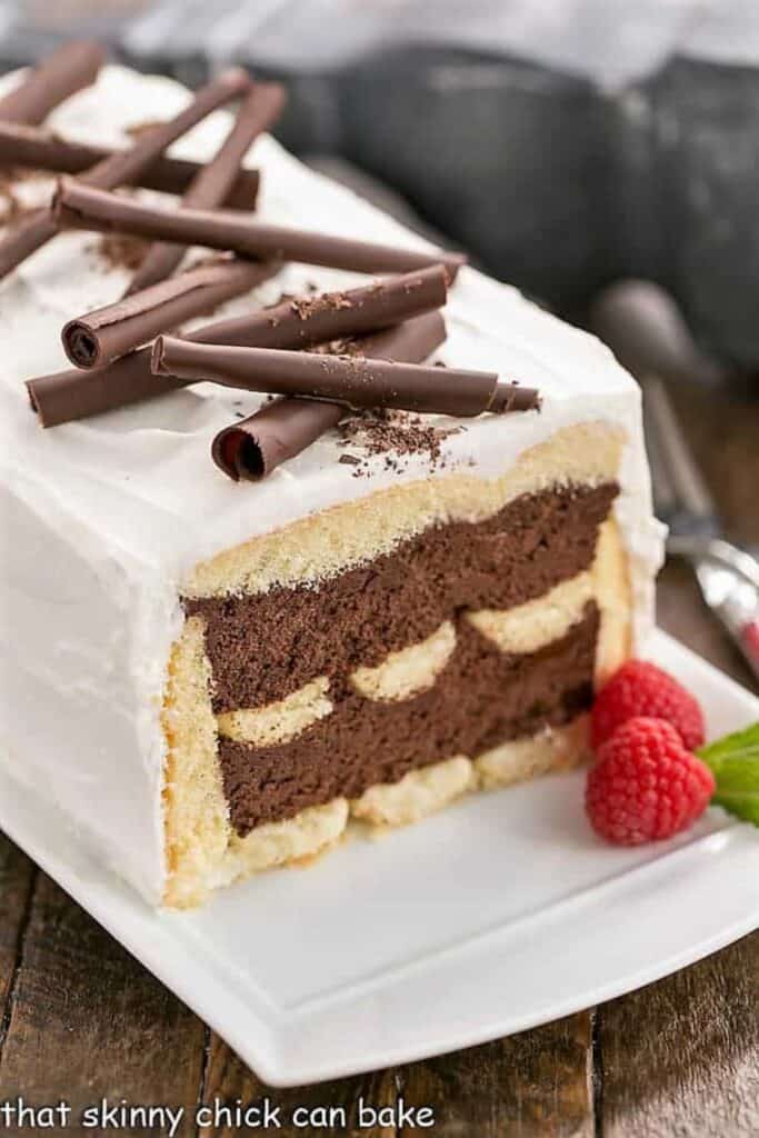A chocolate mousse cake with white icing and shaved chocolate on top with raspberries on a white plate.