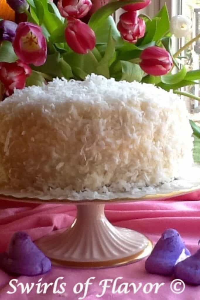 A cocounut lemon layer cake with coconut on the outside on the icing with dark pink tulips in the background.