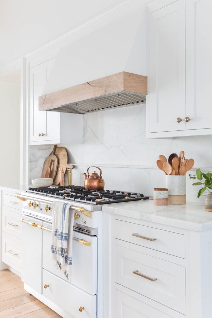 White kitchen with whtie cabinets, white marble counters, gold hardware and a bronze teapot on the stove.