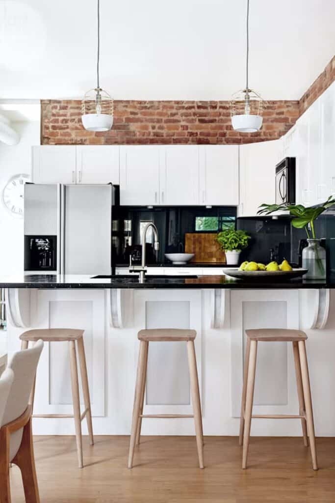A white kitchen with black counters and black backsplash with brick above the cabinets and stainless steel appliances.