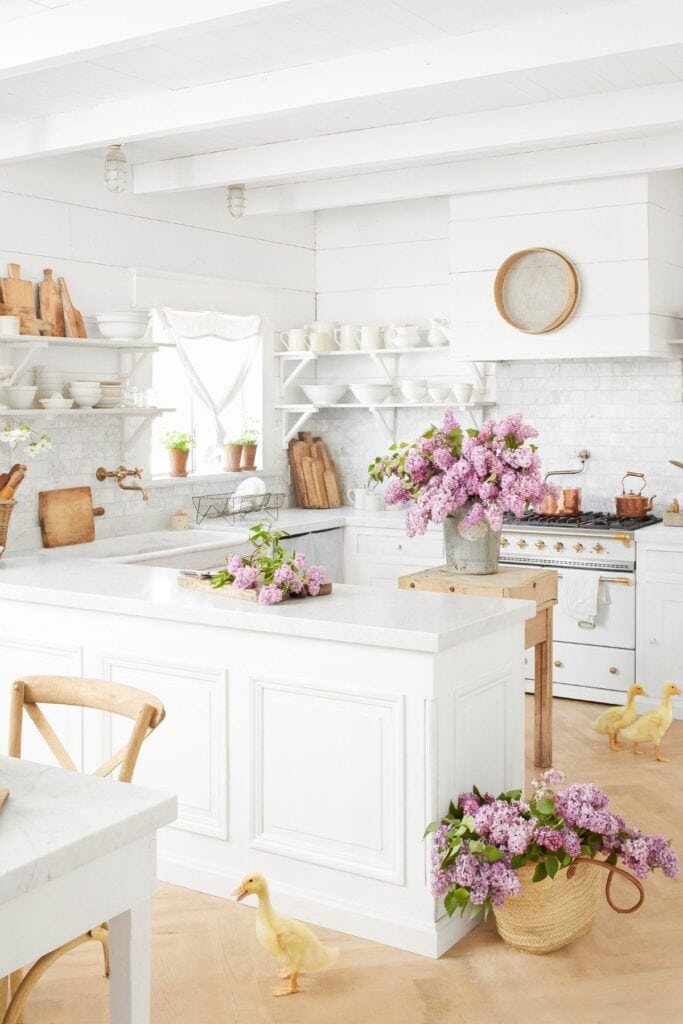 An all white kitchen with shiplap walls and open shelves with white dishes, lots of wooden cutting boards, a wooden island and purple flowers everywhere.