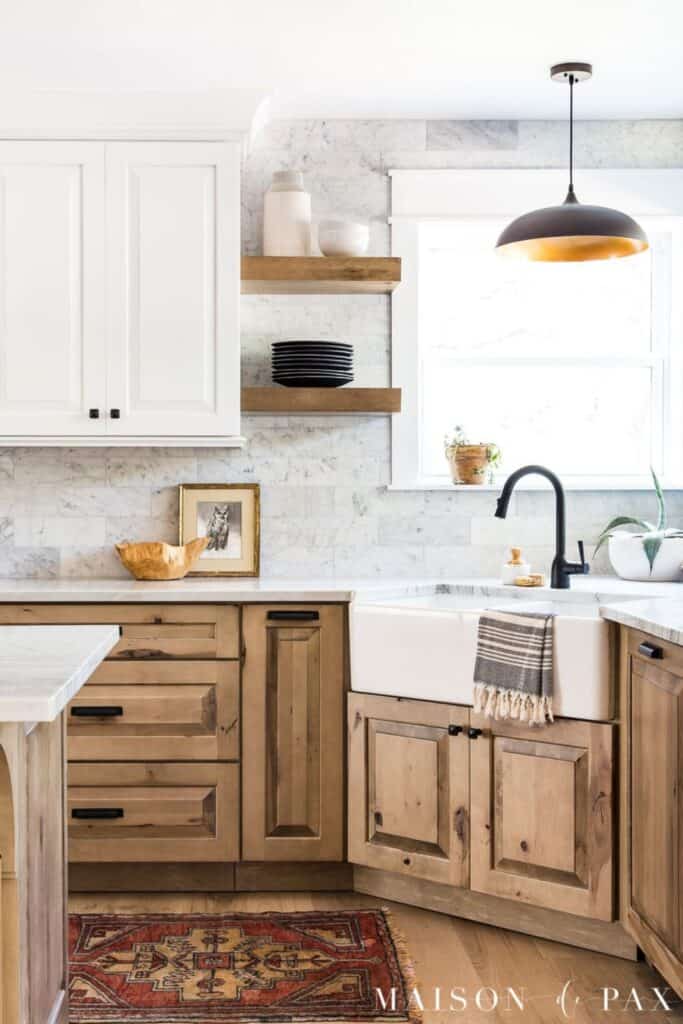 a kitchen with wood lower cabinets and white upper cabinets with a corner sink that has windows above.