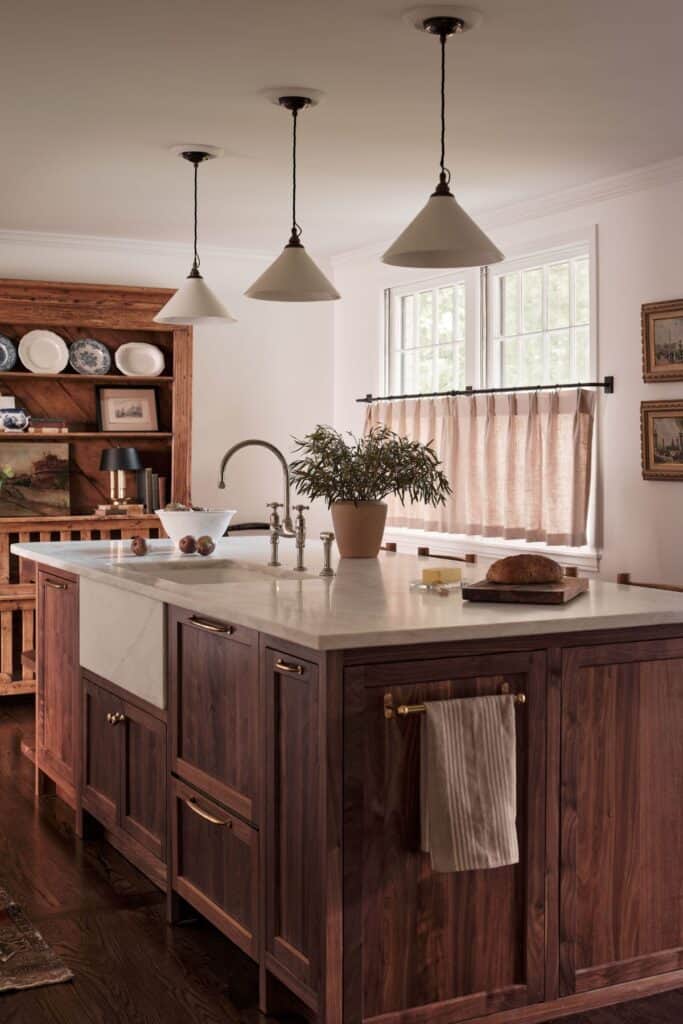 A dark wood kitchen with white counters a farmhouse sink and windows with cafe curtains.